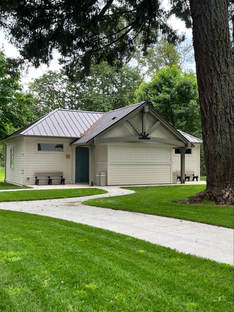 The exterior of a single-story cabin with white shingle and lap siding and a gray metal roof. A concrete walkway leads to the entrance of the cabin. A tree stands in the foreground. Many other trees are visible behind the cabin.