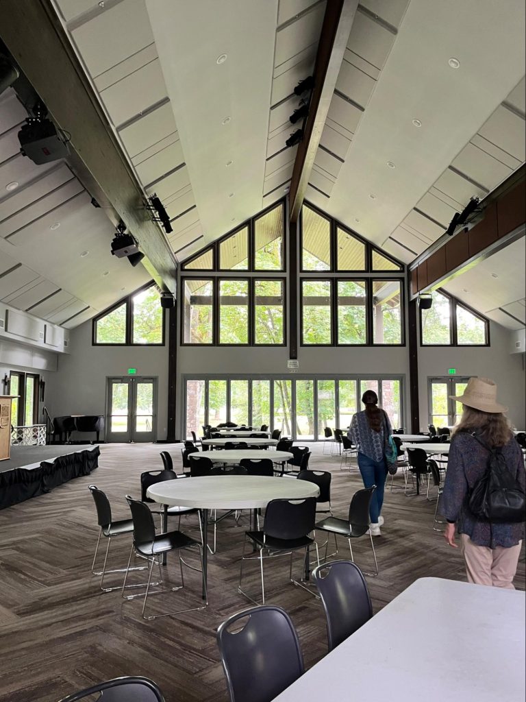 The main hall of the camp. Carpeted and filled with round tables. The back wall of the hall is entirely windows looking out onto natural grounds.